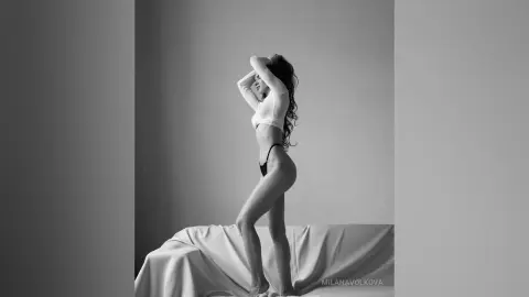 MilanaVolkova poses in profile wearing a white crop top and black panties, showing her bare legs and stomach against a plain background.