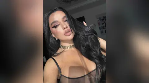 NicoleBella poses with long wavy black hair wearing a black sheer bra and a gold choker necklace.