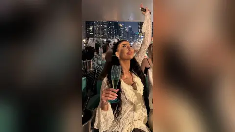Glamorous woman in a white lace blouse holding champagne at a rooftop party with city skyline views at night.