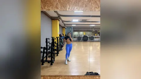 Fit woman in blue leggings and black sports bra posing in a bright modern gym setting.