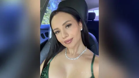 DeviaTelerr poses in a car wearing a green top and a silver necklace, looking at the camera with long dark hair.