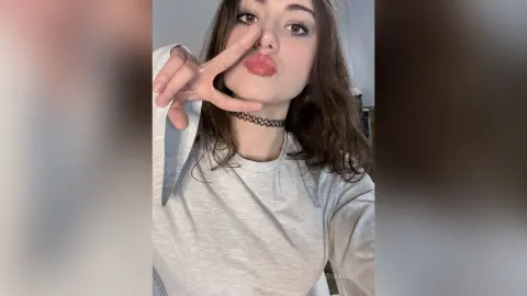 AriaLush pouts her lips and makes a peace sign, wearing a light grey long-sleeve shirt and a black choker.