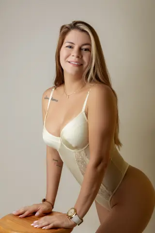 Smiling woman in elegant cream lace bodysuit posing in a bright, minimalist indoor setting.