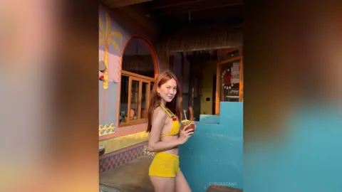 CheCascano poses in a yellow bikini while holding a tropical drink outside a colorful beach hut.