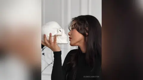 Young woman in a black turtleneck holding a white skull near her face in a minimalist studio setting.