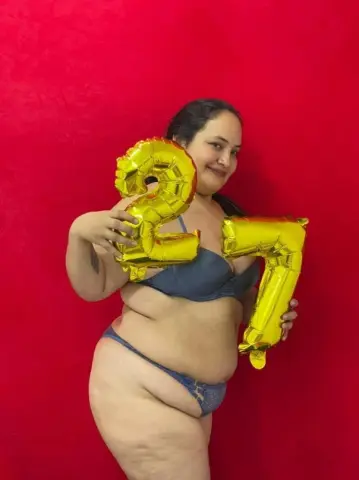 Curvy woman in blue lingerie holding a gold number 2 balloon against a red background for a birthday celebration.