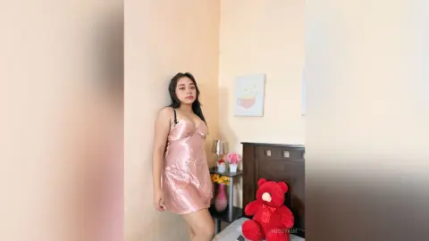 Young woman in a pink satin slip dress posing in a cozy, sunlit bedroom with a red teddy bear.