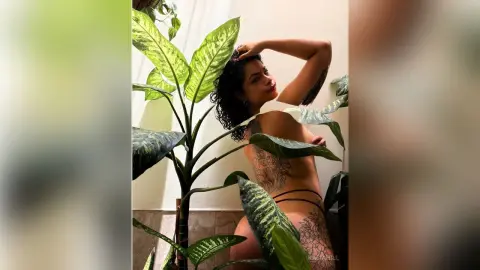 Curvy woman in black bikini posing amidst lush green tropical plants in a bright indoor setting.