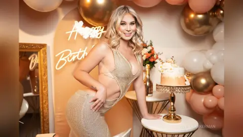 Smiling blonde woman in a sparkling gold dress celebrating a birthday with cake and balloons.