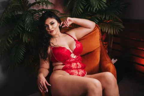 Curvy woman in red lace lingerie relaxing on an orange velvet chair in a lush tropical indoor setting.