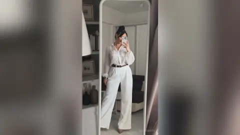 Stylish woman in a white blouse and wide-leg trousers taking a mirror selfie in a modern minimalist bedroom.