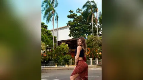 Smiling woman in a black crop top and red patterned skirt posing in a tropical outdoor garden setting.