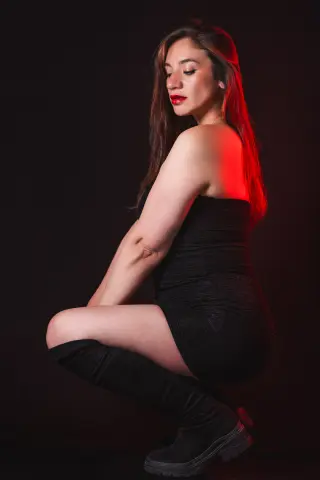 Elegant woman in a black cocktail dress and boots posing against a moody red studio background.
