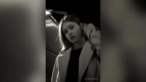 Moody black and white portrait of a young woman in a stylish coat posing outdoors at night.