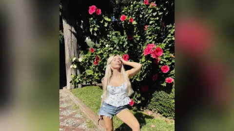 Blonde woman in a floral corset top and denim shorts posing by blooming pink hibiscus flowers in a sunny garden.