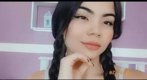 Young woman with dark braided hair and winged eyeliner smiling in a purple bedroom setting.