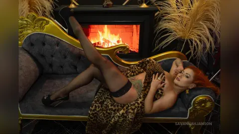 Glamorous redhead in a leopard print robe lounging on a vintage velvet sofa by a warm fireplace.
