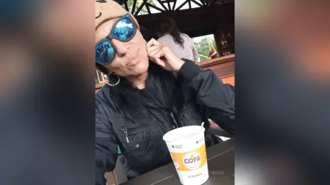 Smiling woman in a black jacket and blue sunglasses relaxing at an outdoor cafe with a coffee cup.