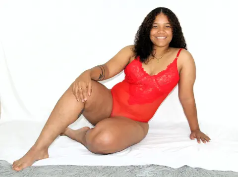 Smiling woman in a red lace bodysuit sitting on a white bed in a bright, minimalist setting.