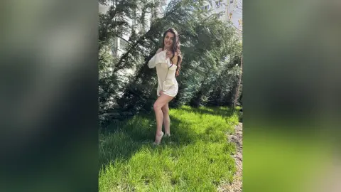 Smiling woman in a white summer outfit standing in a lush green garden under bright sunlight.
