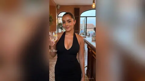 FabianaLima wears a low-cut black halter dress while holding a glass of white wine or cocktail in a bright room.