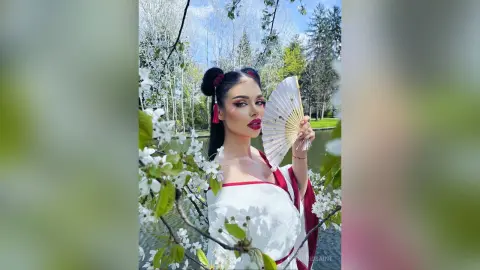 IngridSaint poses in a white and red outfit holding a paper fan amidst blooming white flowers and trees.