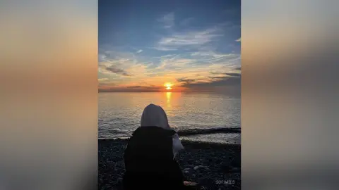 Woman in a white hijab and black outfit sitting on a rocky beach watching a peaceful ocean sunset.