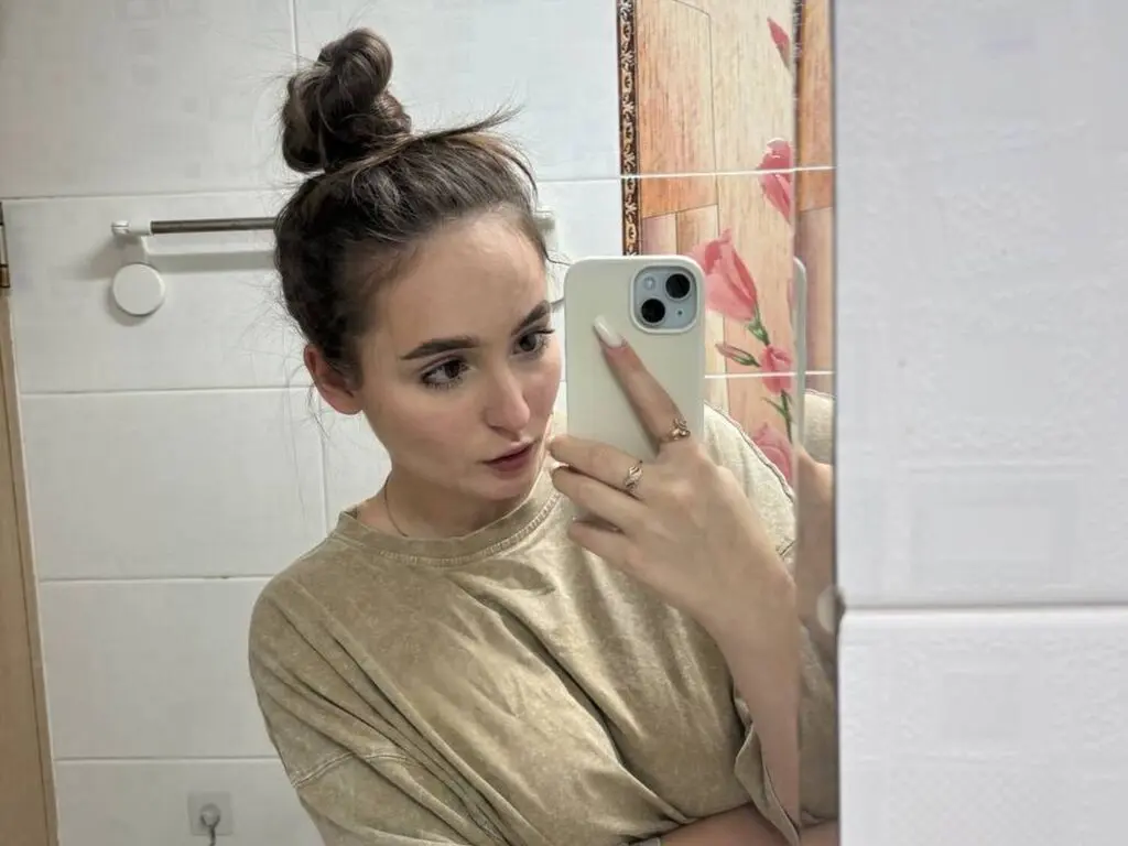 Young woman in a beige casual t-shirt taking a selfie in a bright bathroom mirror.