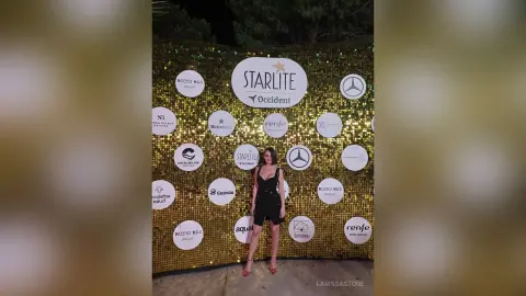 Elegant woman in a black jumpsuit posing at a glamorous Starlite gala event with a gold sequin backdrop.