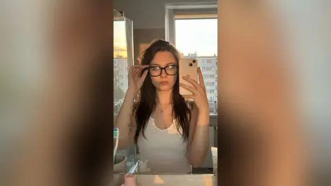 Young woman in a white tank top taking a mirror selfie in a sunlit room during golden hour.