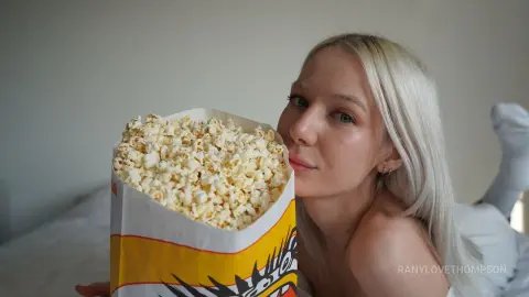 Blonde woman in white loungewear holding a large bag of popcorn in a cozy, bright indoor setting.