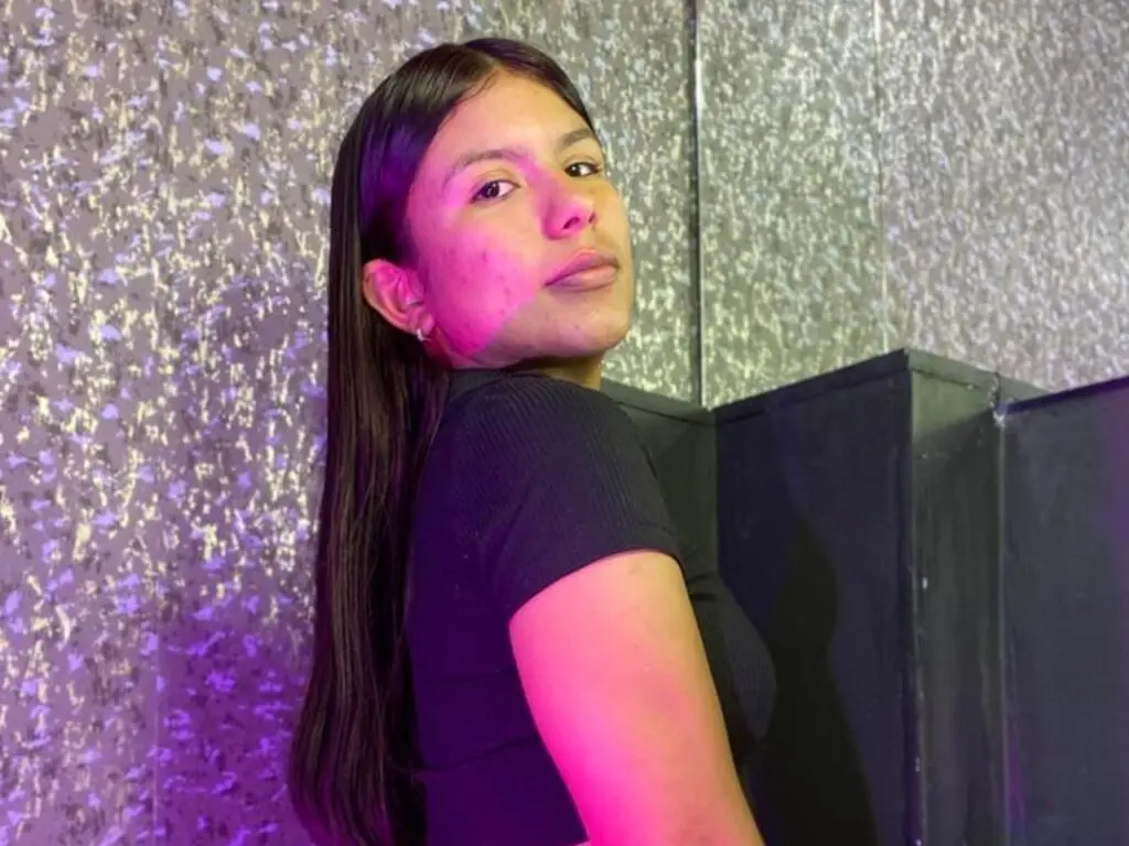 Young woman in a black ribbed top posing against a shimmering silver glitter backdrop in moody purple lighting.