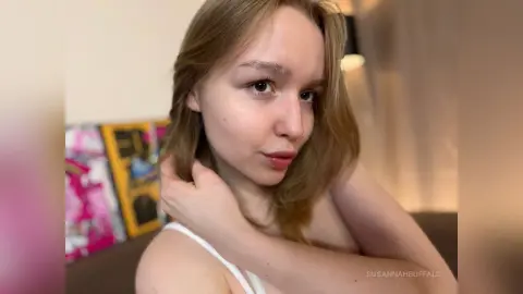 Blonde woman in a white camisole looking thoughtfully in a soft-lit indoor setting.
