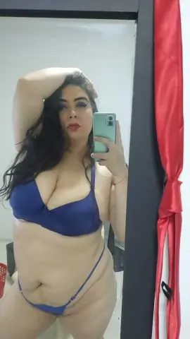 Curvy woman in blue lingerie taking a confident mirror selfie in a bright bedroom setting.
