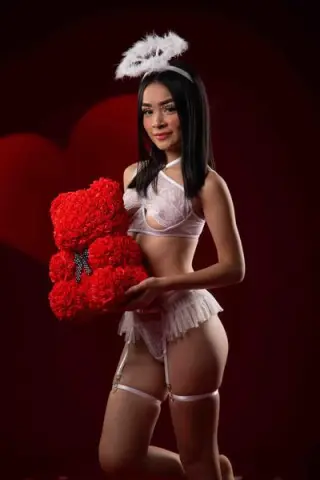 Young woman in white lace lingerie and feathered headband holding a large red rose bouquet against a dark background.