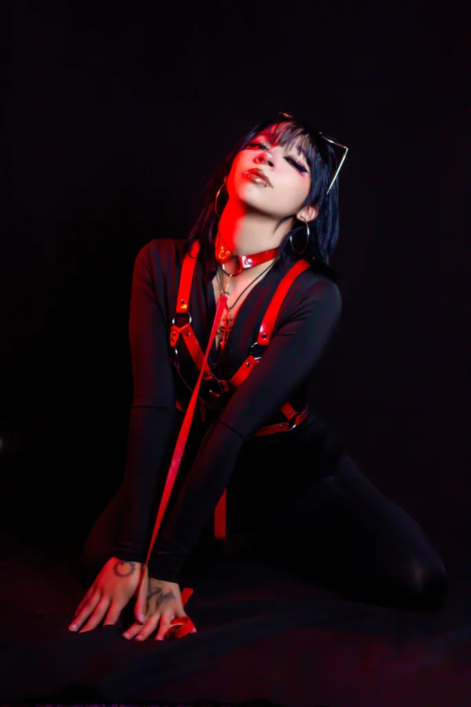 Nina_Ssisy with dark hair and bangs sits in a black chair, wearing a red and black lace corset while holding a red rose to her lips.