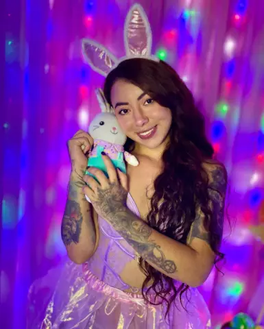 Smiling woman in a pink fairy costume holding a white bunny plushie against a colorful disco light background.
