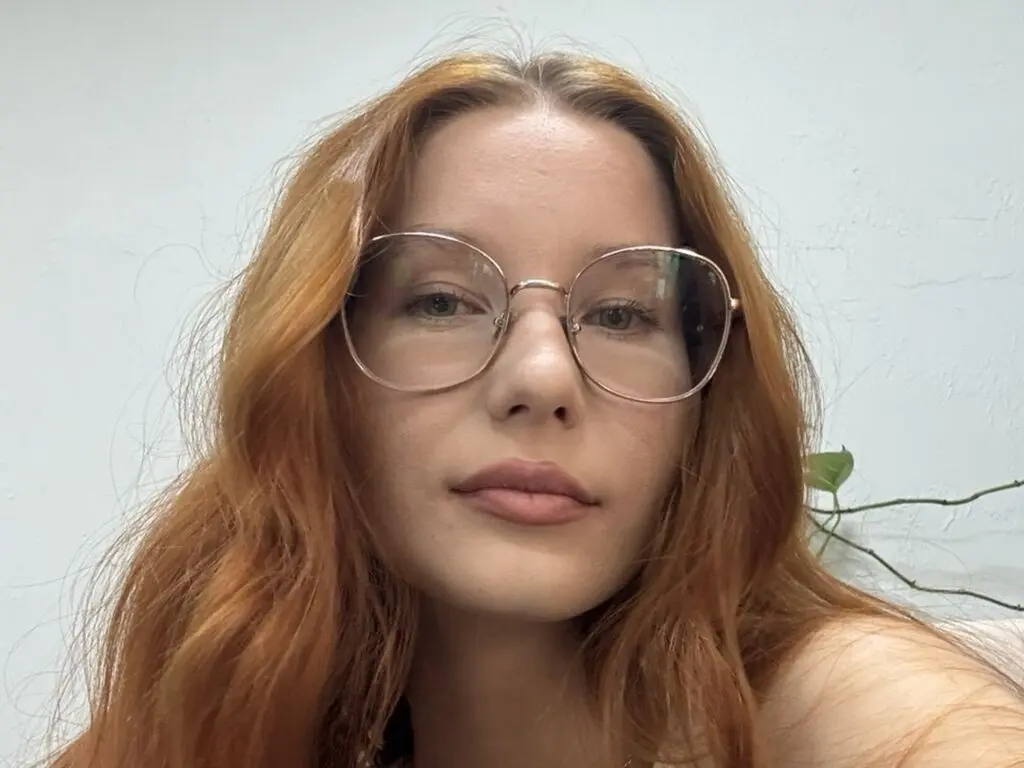 Redheaded woman with glasses looking calmly at the camera in a bright indoor setting.