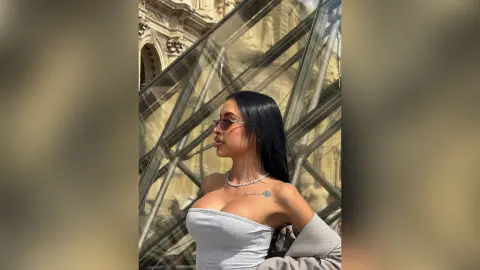 Stylish woman in a grey tube top and sunglasses posing by the glass architecture of the Louvre in Paris.