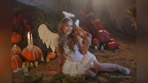 StellaFletcher wears a white lace dress and white wings while sitting on sand surrounded by pumpkins and lit candles.
