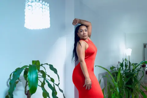 Elegant woman in a red sleeveless dress posing in a bright modern room with green houseplants.