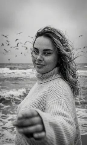 Smiling woman in a cozy knit turtleneck sweater standing on a windy beach under a cloudy sky.