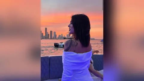 Woman in white off-the-shoulder dress enjoying a sunset boat ride on the ocean with a city skyline view.