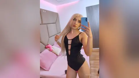 Blonde woman in a black cutout bodysuit taking a mirror selfie in a bedroom with pink mood lighting.