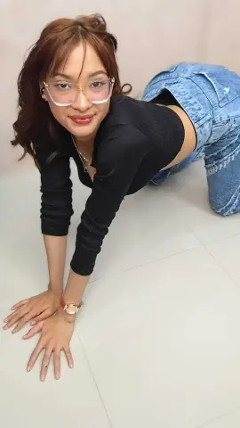 Smiling young woman in a black long-sleeve top and blue jeans posing on a white floor.