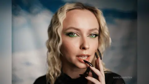 Blonde woman with green eyeliner and long black nails posing against a soft blue sky background.