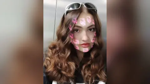 VickyToure poses with long wavy brown hair and sunglasses on her head, looking directly at the camera with pink heart-shaped filters on her face.