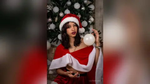 Smiling woman in a red and white Santa outfit holding a white ornament by a decorated Christmas tree.