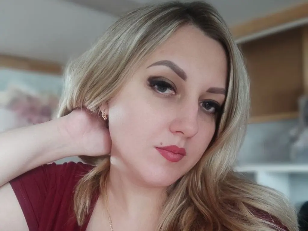 MariaPixie poses with her hand behind her neck, showing her blonde hair and red lipstick in a close-up shot.