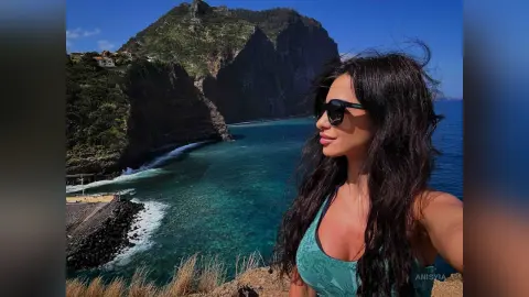 Smiling woman in a green tank top enjoying a sunny coastal view overlooking turquoise ocean waves and cliffs.